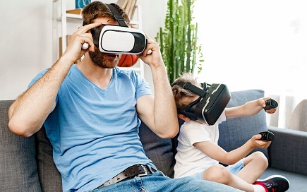 Auf einer Couch sitzen Vater und Sohn. Beide tragen eine virtuelle Brille. Der Sohn hat in jeder Hand einen Joystick.